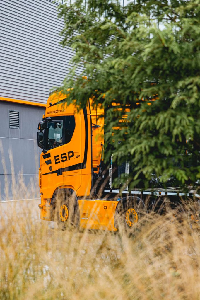 Gele vrachtwagen achter boom bij industrieel gebouw